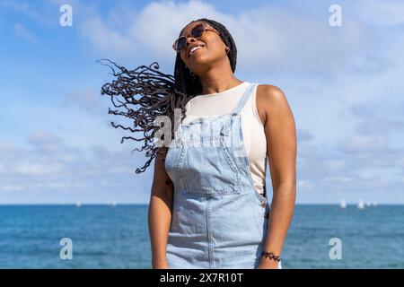 Une jeune femme heureuse en salopette en denim sourit avec la mer derrière elle lors d'une journée ensoleillée de Barcelone Banque D'Images