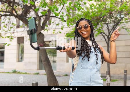 Heureuse jeune femme prenant un selfie avec un smartphone sur un bâton de selfie, pointant et souriant en plein air lors d'une journée d'été ensoleillée à Barcelone Banque D'Images