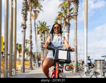 Une femme joyeuse faisant du vélo sous le soleil d'été le long d'un boulevard bordé de palmiers à Barcelone, dégageant des vibrations insouciantes et un style de vie urbain Banque D'Images
