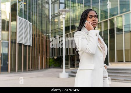 Une femme d'affaires afro-américaine s'engage dans une conversation téléphonique sérieuse tout en se tenant debout devant un immeuble de bureaux en verre moderne, vêtue d'un style Banque D'Images