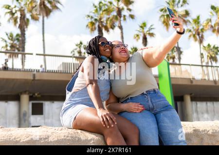 Deux amis partagent un joyeux moment selfie sous le soleil de Barcelone, entourés de palmiers et d’architecture locale Banque D'Images