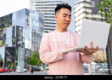 Un jeune homme asiatique dans une tenue rose décontractée tient et utilise intentionnellement un ordinateur portable par une journée ensoleillée, debout devant les bâtiments modernes de la ville. Banque D'Images