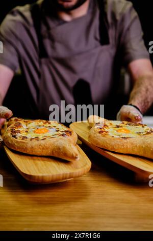 Un chef, partiellement visible, sert du khachapuri géorgien fraîchement cuit, un pain fourré au fromage avec un œuf ensoleillé au centre, présenté sur des planches de bois Banque D'Images