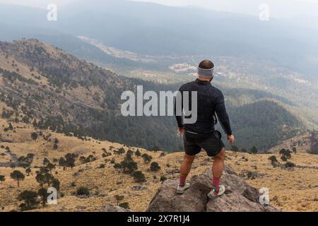 Un randonneur masculin se tient sur un pic rocheux, contemplant le vaste terrain montagneux dans le cadre de sa séance d'entraînement en plein air Banque D'Images