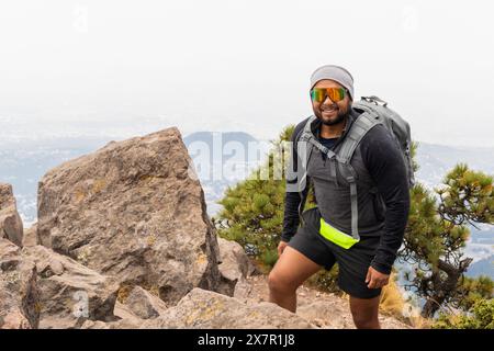 Un randonneur joyeux avec un sac à dos s'arrête sur un sommet de montagne, surplombant un paysage brumeux tout en s'entraînant pour de futurs treks Banque D'Images