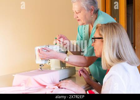 Deux femmes engagées dans un projet de couture avec l'une d'entre elles apprennent à l'autre comment utiliser une machine à coudre, indiquant un environnement de leçon ou d'atelier Banque D'Images