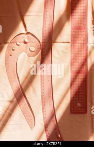 Des outils de couture tels qu'une courbe française et un ruban à mesurer projettent des ombres douces sur un tissu rose, créant une scène artistique d'atelier de couture Banque D'Images