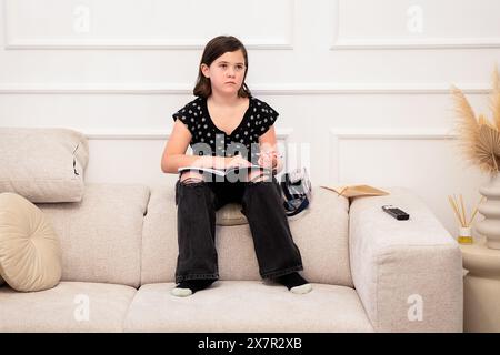 Une jeune fille de preteen est assise confortablement sur un canapé beige à la maison, engrossée dans un livre avec un look pensif, portant des vêtements décontractés, et regardant loin de la CA Banque D'Images