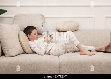 Un jeune garçon se prélasse sur un canapé, concentré sur son smartphone, vêtu d’un pyjama blanc à pois noirs. Banque D'Images