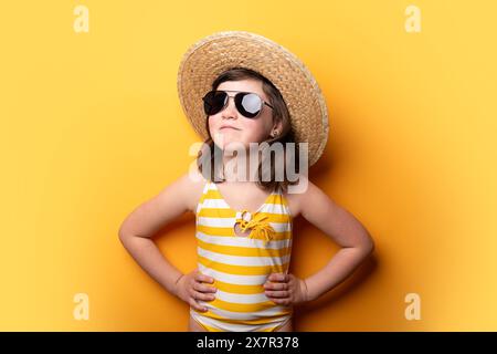 Une jeune fille nageuse vêtue d'un maillot de bain rayé jaune et blanc, arborant un chapeau de paille et des lunettes de soleil élégantes, se tient confiante contre un jaune vif Banque D'Images