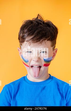 Un jeune garçon ludique portant une chemise bleu vif sort sa langue, affichant de la peinture arc-en-ciel sur ses joues contre un dos orange vif Banque D'Images