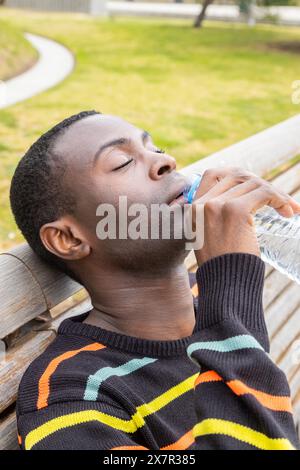 Un jeune homme est assis sur un banc de parc, buvant de l'eau dans une bouteille, combattant peut-être les symptômes de l'hypoglycémie Banque D'Images