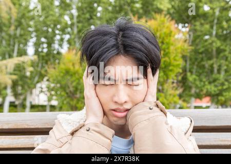 Un jeune homme asiatique est assis sur un banc de parc serrant sa tête dans l'inconfort, dépeignant une scène liée à des symptômes d'hypoglycémie Banque D'Images