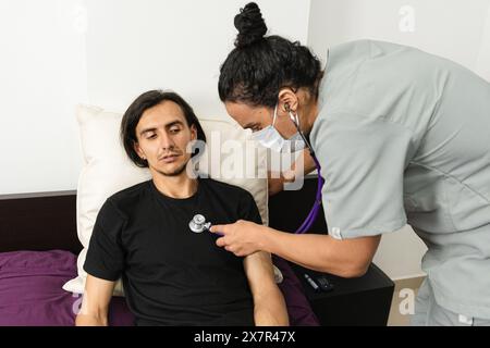 Une infirmière en uniforme gris utilise un stéthoscope pour vérifier les symptômes de l'hypoglycémie sur un patient masculin allongé sur un lit dans un cadre familial Banque D'Images