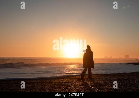 Une image sereine capturant la silhouette d'un individu marchant le long d'une plage islandaise, avec le soleil se levant brillamment sur l'horizon et les vagues Banque D'Images