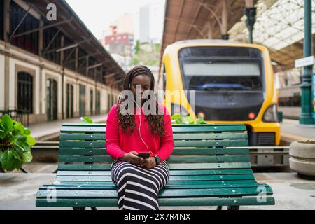Une jeune femme noire est prise dans son smartphone alors qu'elle est assise sur un banc vert à une gare, avec un train jaune en arrière-plan Banque D'Images