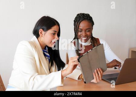 Deux femmes diverses examinent les données sur une tablette numérique tout en travaillant ensemble sur un projet d'entreprise dans un cadre confortable à la maison, mettant en valeur le travail d'équipe et la technol Banque D'Images