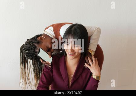 Deux femmes joyeuses partageant un rire lors d'une pause détente à la maison, l'une tenant un smartphone, toutes deux vêtues d'une tenue chic et décontractée Banque D'Images