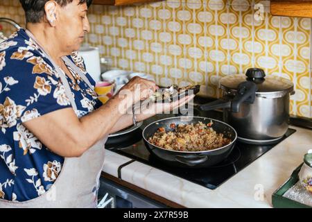 Une femme âgée dans un tablier floral cuisine et assaisonne des aliments dans une poêle à frire dans sa cuisine à la maison, démontrant la vie quotidienne et les compétences culinaires Banque D'Images