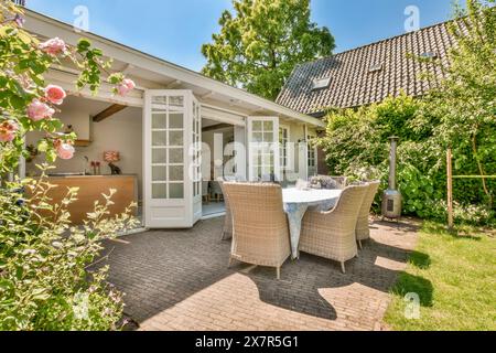 Une salle à manger extérieure accueillante avec une table et des chaises élégantes sur un patio pavé, entouré de verdure luxuriante et de fleurs roses dans une maison confortable sur Banque D'Images