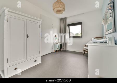 Une chambre minimaliste avec une grande armoire blanche, une élégante commode et un sol gris doux la lumière naturelle traverse une fenêtre, complétée par une fenêtre Banque D'Images