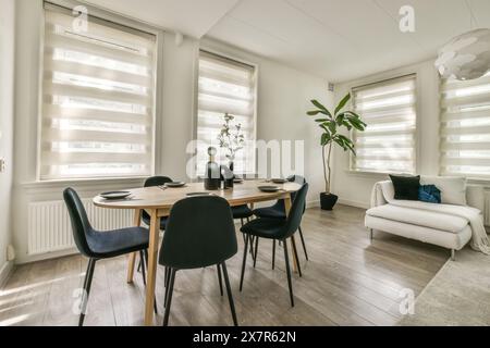Un espace salle à manger moderne avec un élégant ensemble de salle à manger noir, un canapé blanc confortable et des fenêtres habillées de stores blancs, le tout éclairé par la lumière naturelle Banque D'Images
