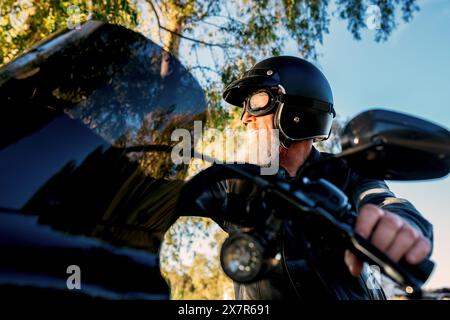 D'en bas un motocycliste masculin âgé avec un casque noir et des lunettes est à cheval et regarde loin de la caméra, montrant un sentiment de liberté et d'adv Banque D'Images