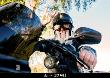 D'en bas homme âgé joyeux avec une barbe portant un casque donne un signe de paix tout en conduisant une moto, en regardant la caméra. Banque D'Images