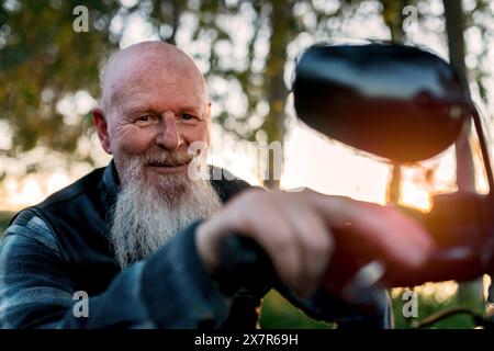 Un vieil homme joyeux avec une barbe blanche et une expression souriante est vu ajuster le rétroviseur latéral d'une moto pendant le coucher du soleil. Il regarde Cam Banque D'Images