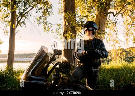 Motocycliste masculin senior en cuir se reposant à côté de son vélo, profitant du coucher du soleil. Banque D'Images