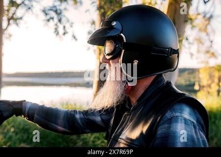 Profil de vue latérale d'un motocycliste masculin senior avec une barbe portant un casque et des lunettes regardant loin de la caméra. Banque D'Images
