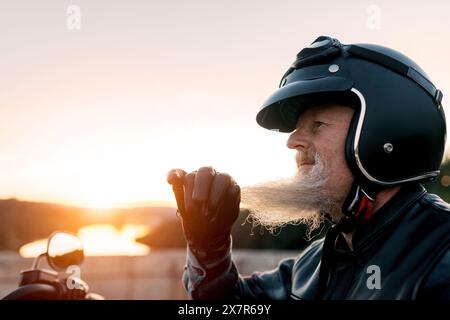 Vue latérale d'un motocycliste masculin âgé dans un casque regarde loin tout en savourant la liberté d'une promenade panoramique au coucher du soleil, exprimant l'aventure et la sérénité R Banque D'Images