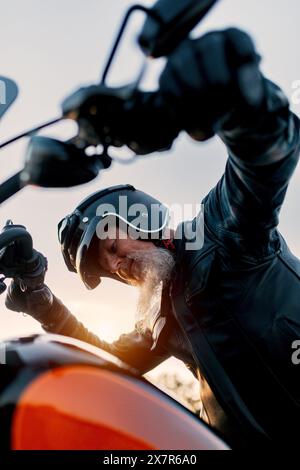D'en bas, un motard masculin aîné avec une longue barbe ajuste son casque avant une promenade au coucher du soleil, dégageant un sentiment d'aventure et d'expérience. Il l'est Banque D'Images