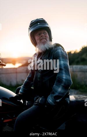 Un motard masculin mature avec une barbe blanche, portant un casque et une veste en cuir, est assis sur une moto, regardant loin pendant un coucher de soleil. Banque D'Images