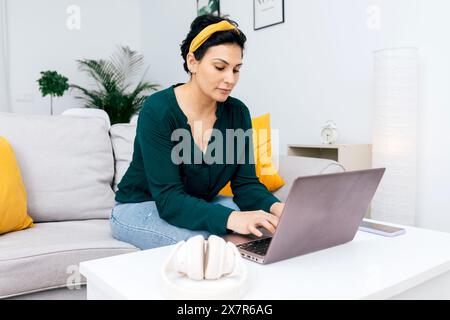 Une femme concentrée dans une blouse vert foncé s'engage avec son ordinateur portable tout en s'asseyant confortablement sur un canapé blanc dans un salon lumineux, orné de M. Banque D'Images