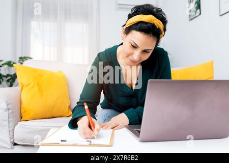 Une femme vêtue d'une blouse verte travaille à la maison, écrivant dans son cahier tout en utilisant son ordinateur portable son bureau à domicile détendu avec cushi jaune Banque D'Images