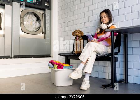 Une jeune fille de preteen est assise sur un banc avec son chien de compagnie, regardant son smartphone, attendant que la lessive soit terminée. Banque D'Images