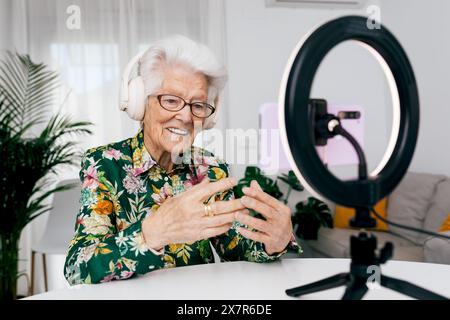 Podcaster féminin senior utilisant des écouteurs et une lumière annulaire, engageant son public à travers un smartphone, affichant vitalité et enthousiasme dans un ho Banque D'Images