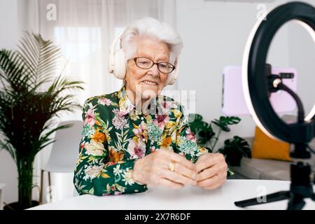 Joyeuse dame âgée dans une chemise florale utilise des écouteurs et enregistre un podcast ou une vidéo, interagissant avec son public par le biais des médias sociaux Banque D'Images