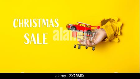 Une main tenant une voiture jouet dans un panier avec les mots soldes de Noël écrit ci-dessous. L'image a une ambiance ludique et festive, suggérant que le Banque D'Images
