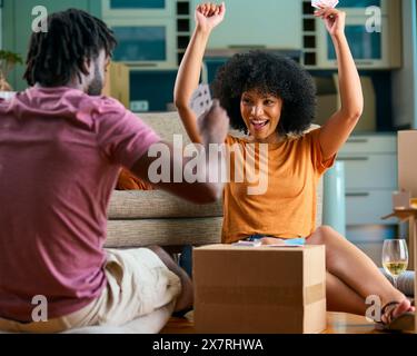 Jeune couple jouant des cartes dans la nouvelle maison le jour de déménagement boire du vin Banque D'Images