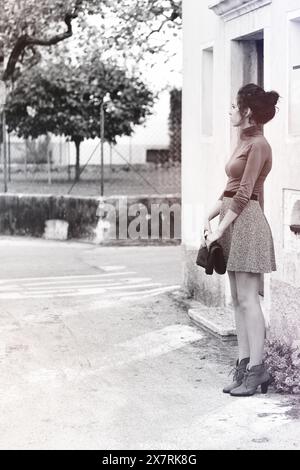 jeune femme, dans un col roulé brun et une jupe grise, se tient près d'un bâtiment. Elle regarde de côté, tenant une pochette. L'image en noir et blanc montre un tre Banque D'Images