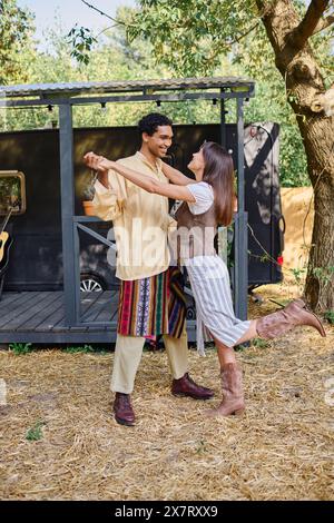Un homme et une femme de différentes races se balancent gracieusement, dansant devant leur charmante caravane de camping-car dans un cadre naturel pittoresque. Banque D'Images