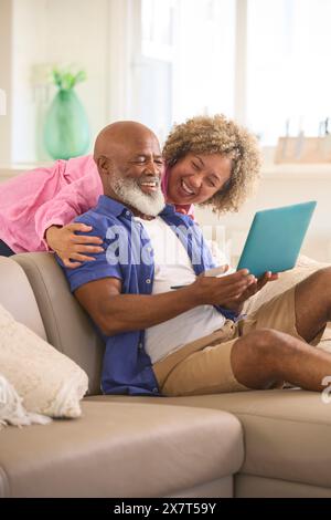 Couple mature à la maison avec femme embrassant et homme surprenant à l'aide d'ordinateur portable assis sur canapé Banque D'Images
