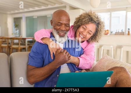 Couple mature à la maison avec femme embrassant et homme surprenant à l'aide d'ordinateur portable assis sur canapé Banque D'Images