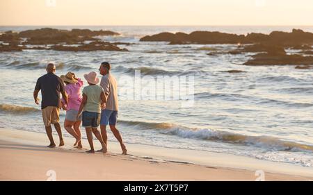 Vue arrière d'un couple d'âge mûr avec des amis en vacances marchant le long de la plage Shoreline au coucher du soleil Banque D'Images