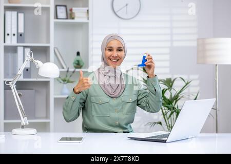 Femme confiante en hijab travaillant à domicile, montrant l'inhalateur d'asthme et donnant le pouce vers le haut geste, concept de santé et de travail à distance Banque D'Images