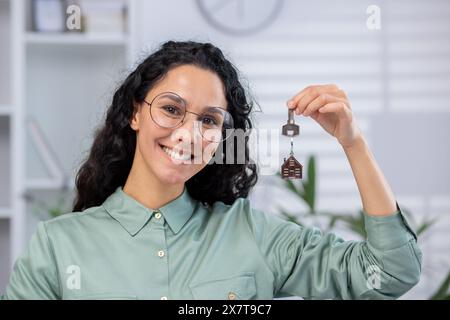 Femme souriante portant des lunettes et chemise verte tenant la clé de la maison avec porte-clés dans un cadre de bureau à domicile. Banque D'Images