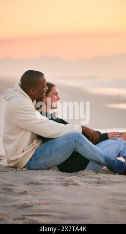 Jeune couple amoureux habillé de façon décontractée assis sur Beach Shoreline embrassant au lever du soleil Banque D'Images