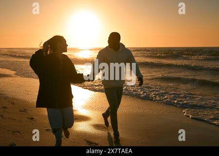 Jeune couple amoureux habillé de façon décontractée courant le long de Beach Shoreline à travers les vagues au lever du soleil Banque D'Images